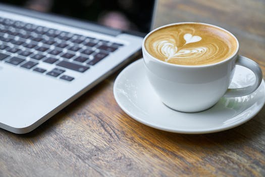 pexels-photo-414630-414630 A cup of latte with intricate art beside a laptop on a wooden table, depicting a cozy work setting.